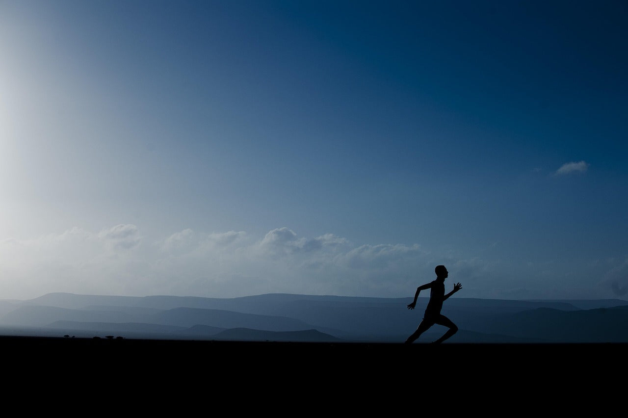 Silhouette d’un coureur en plein effort sur une route au lever du soleil, avec un ciel bleu et des montagnes en arrière-plan.
